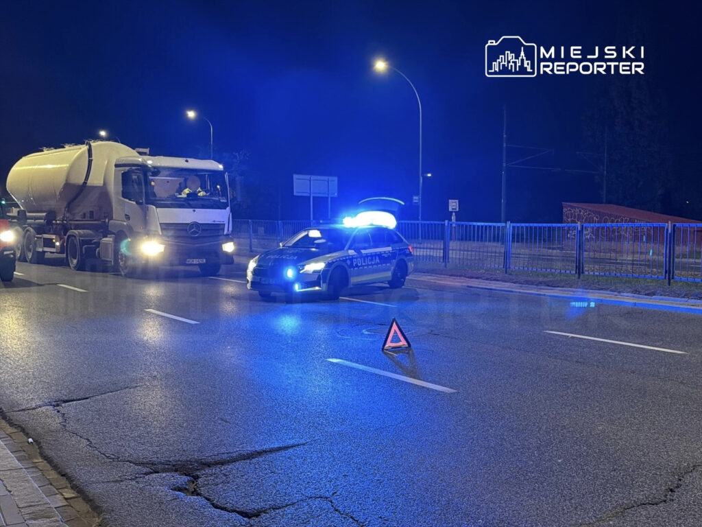 Policyjny radiowóz z włączonymi sygnałami świetlnymi blokuje pas ruchu, obok zatrzymany ciężarowy pojazd.