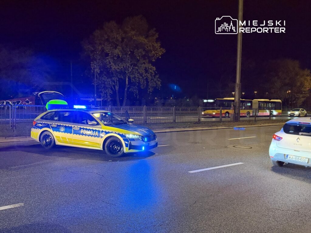 Policyjny radiowóz z niebieskimi światłami stoi na jezdni, obok otwarty bagażnik, w tle przejeżdżający autobus.