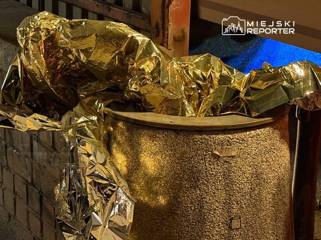 Złota folia owija przedmiot umieszczony na okrągłej, teksturowanej powierzchni w miejskim otoczeniu.