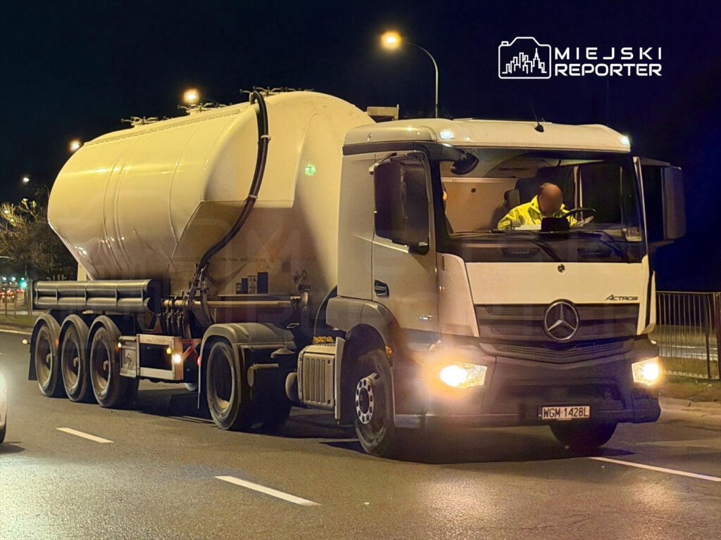 Ciężarówka z białym zbiornikiem porusza się po ulicy nocą, kierowca w żółtej kurtce obsługuje pojazd.