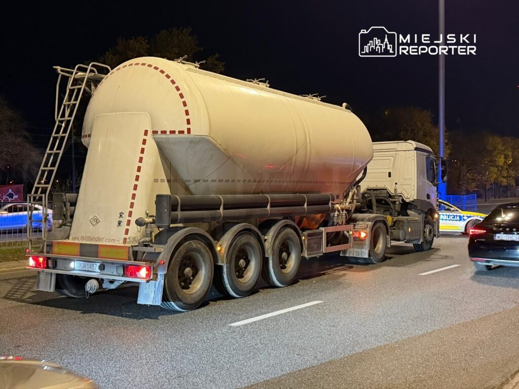 Ciężarówka cysternowa zatrzymana na jezdni, obok policyjny radiowóz z włączonymi sygnałami świetlnymi.