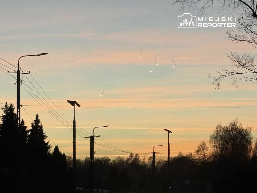Na niebie widać jasne smugi światła, a w tle rozciąga się pastelowy zachód słońca. Ulicę otaczają wysokie latarnie i drzewa.