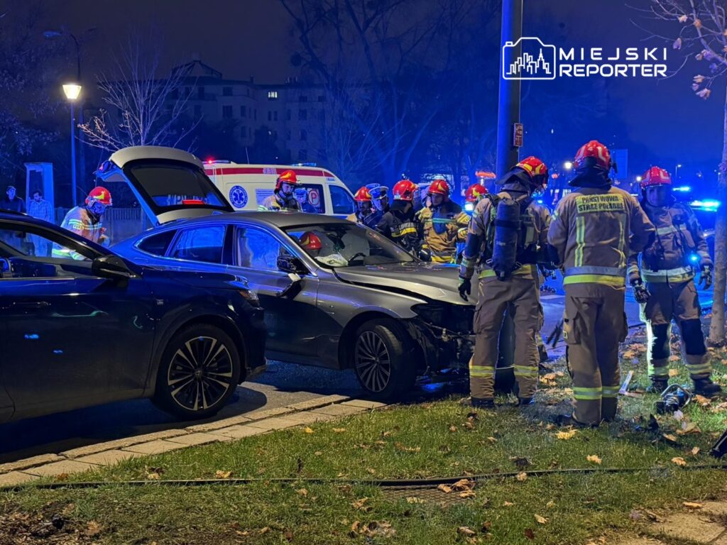 Grupa strażaków w hełmach i mundurach pracuje przy uszkodzonym pojeździe na oświetlonej ulicy w nocy.