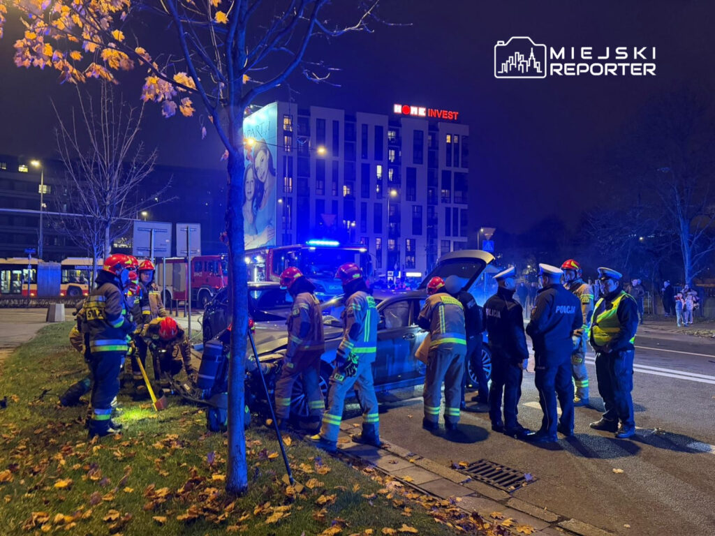Grupa strażaków i policjantów pracuje nad usunięciem pojazdu z rowu na oświetlonej ulicy w nocy.