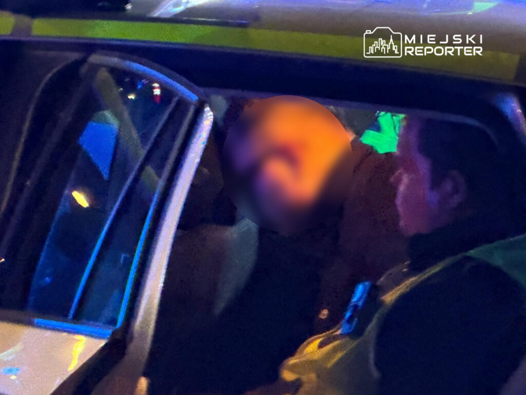 Mężczyzna siedzi na tylnym siedzeniu radiowozu, a policjant obserwuje sytuację w ciemnym otoczeniu.