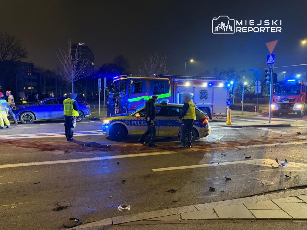 Grupa policjantów i strażaków prowadzi działania na miejscu wypadku drogowego, obok uszkodzonego samochodu.