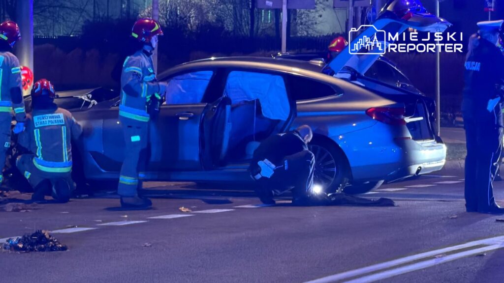 Strażacy w mundurach przeszukują wrak samochodu osobowego na drodze, przy włączonych sygnałach świetlnych.