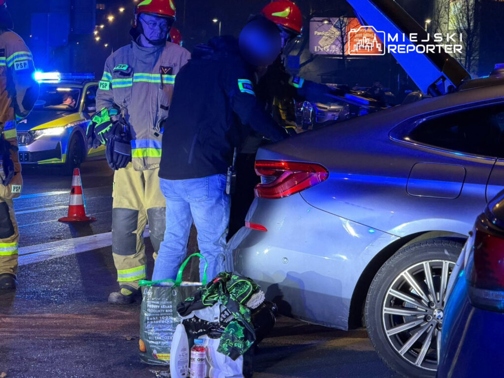 Grupa strażaków w mundurach interweniuje przy uszkodzonym samochodzie na oświetlonej ulicy.