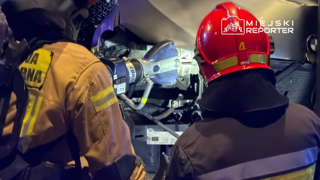 Dwóch strażaków w hełmach pracuje nad usunięciem przeszkody w pojeździe, używając narzędzi hydraulicznych.