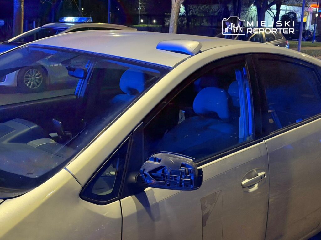 W nocy, biały samochód z uszkodzonym lusterkiem stoi na ulicy, obok widoczne niebieskie światła policyjne.