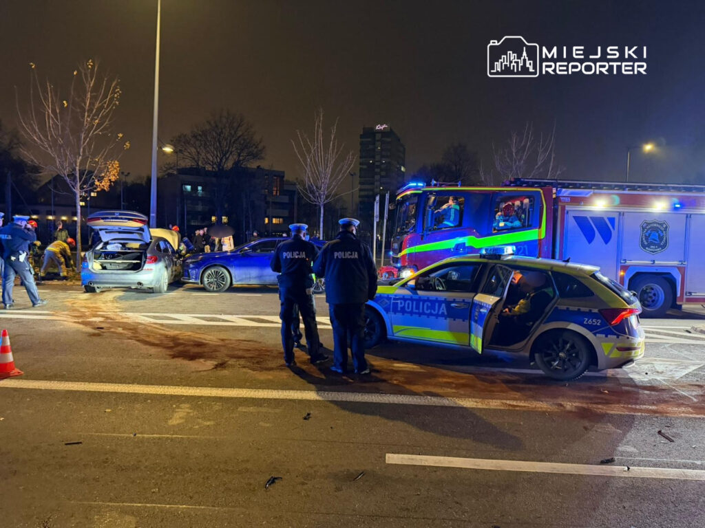 Grupa policjantów i strażaków pracuje przy wypadku drogowym, obok uszkodzonych samochodów i pojazdu strażackiego.