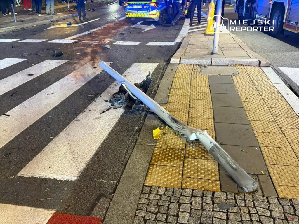 Złamana latarnia uliczna leży na pasach dla pieszych, a w tle widać radiowóz i grupę policjantów.