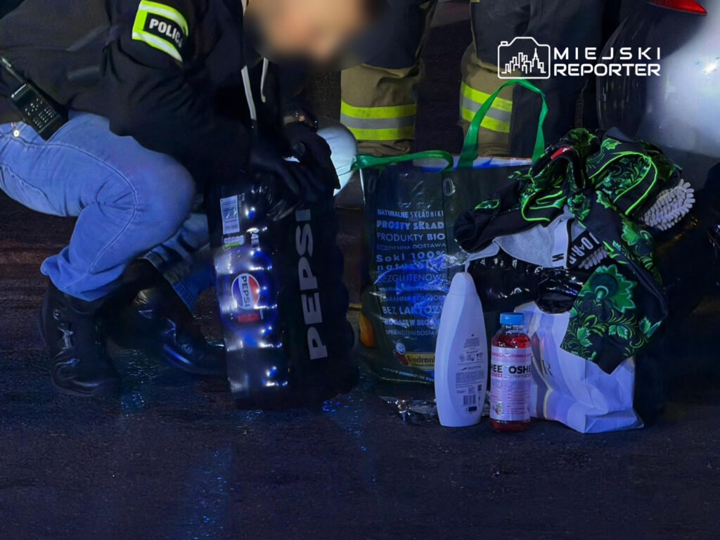 Policjant w mundurze pochyla się nad torbą, obok której leżą butelki napoju oraz kosmetyki na mokrej nawierzchni.