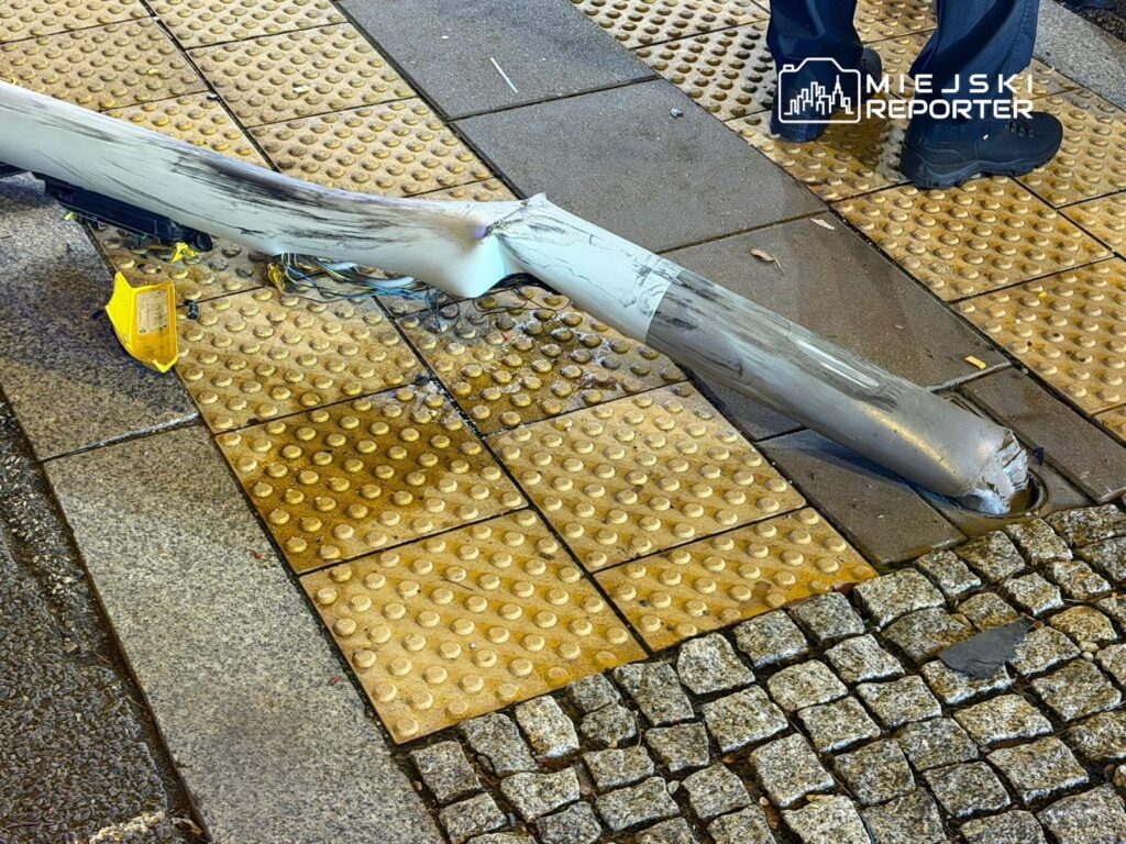 Złamany słup oświetleniowy leży na chodniku, obok widoczne są uszkodzone przewody i żółta tablica ostrzegawcza.