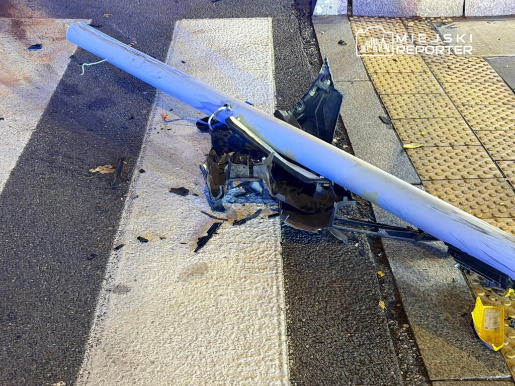 Złamany słup oświetleniowy leży na pasach, obok uszkodzony fragment pojazdu oraz resztki plastikowe.