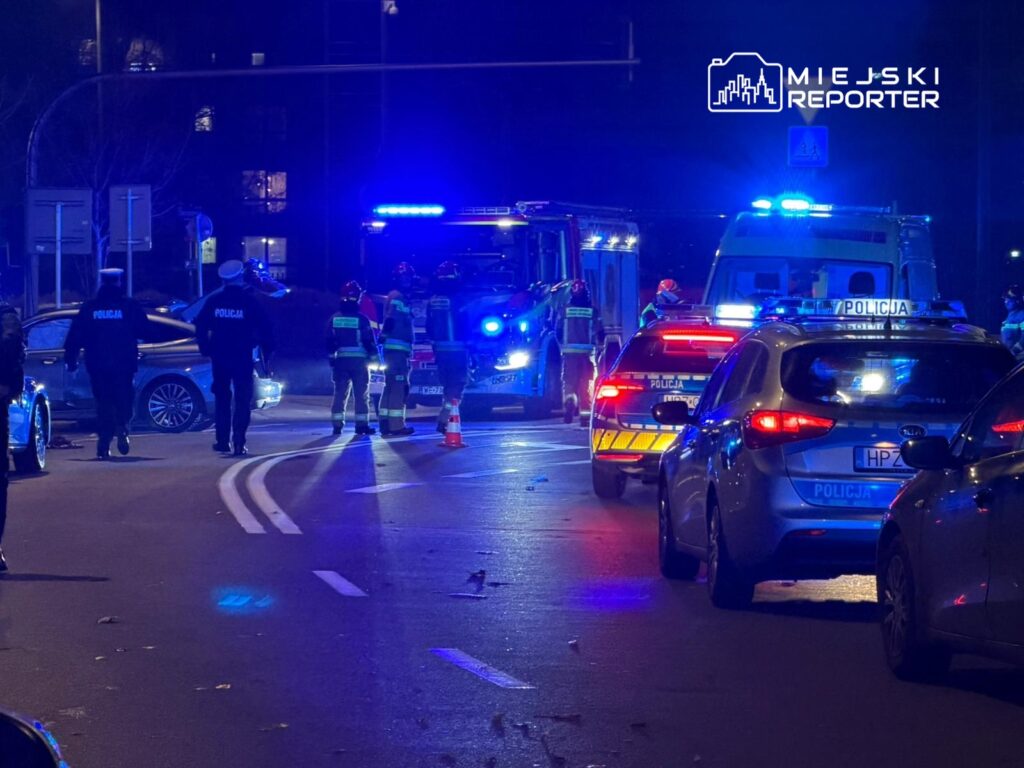 Grupa policjantów i strażaków pracuje na miejscu wypadku drogowego, w tle widać radiowozy z włączonymi sygnałami.