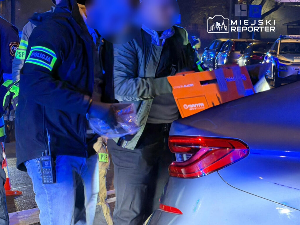 Funkcjonariusze policji przeszukują bagażnik samochodu, trzymając pomarańczowy karton w nocy na oświetlonej ulicy.