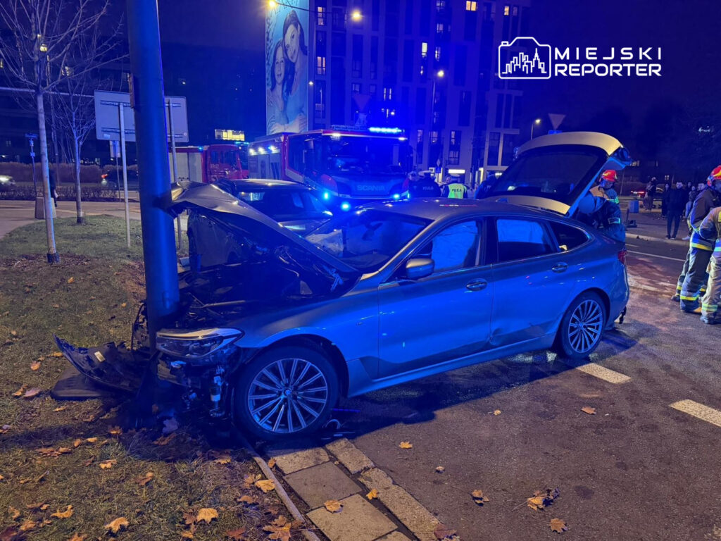 Niebieski samochód osobowy z uszkodzoną przednią częścią uderzył w słup, obok stoją strażacy i policjanci.