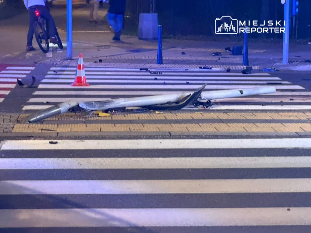 Złamany słup oświetleniowy leży na pasach dla pieszych, obok pomarańczowy pachołek. W tle widać rowerzystę.