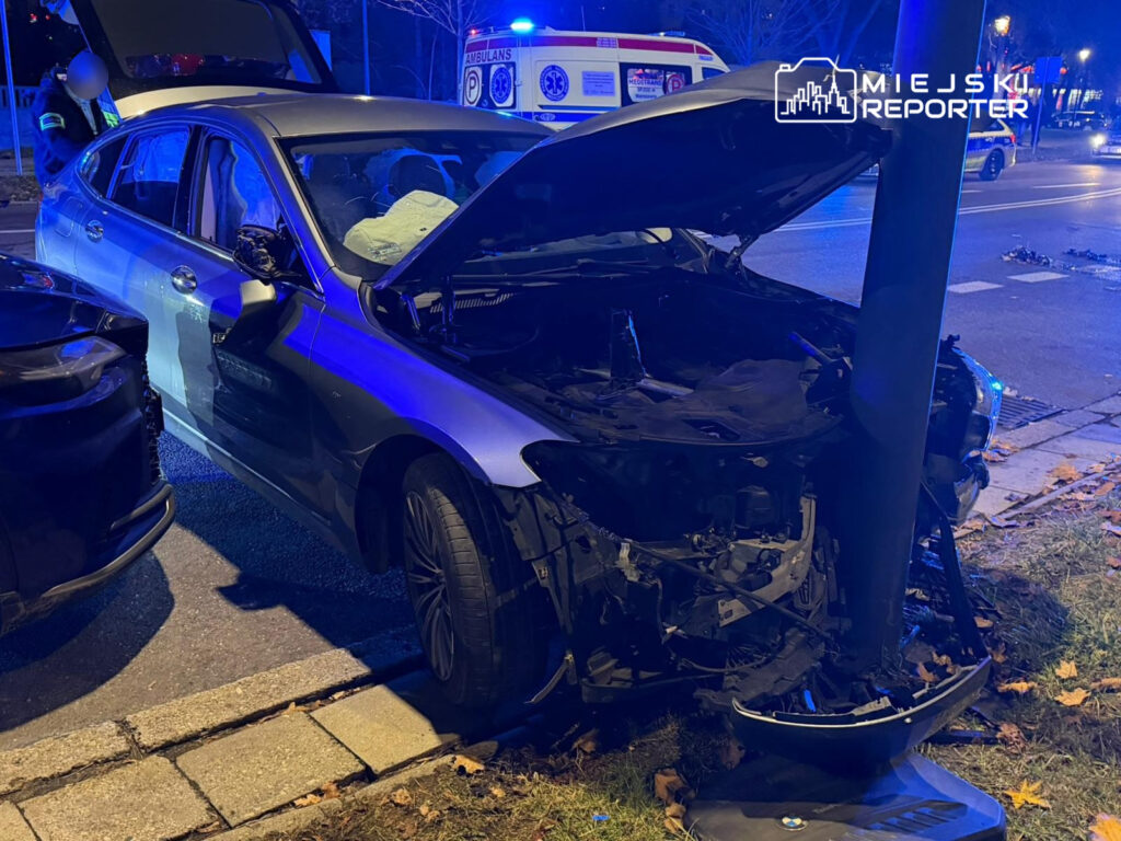 Uszkodzony samochód osobowy z otwartą maską, stojący przy lampie ulicznej, w tle widać pojazd ratunkowy.