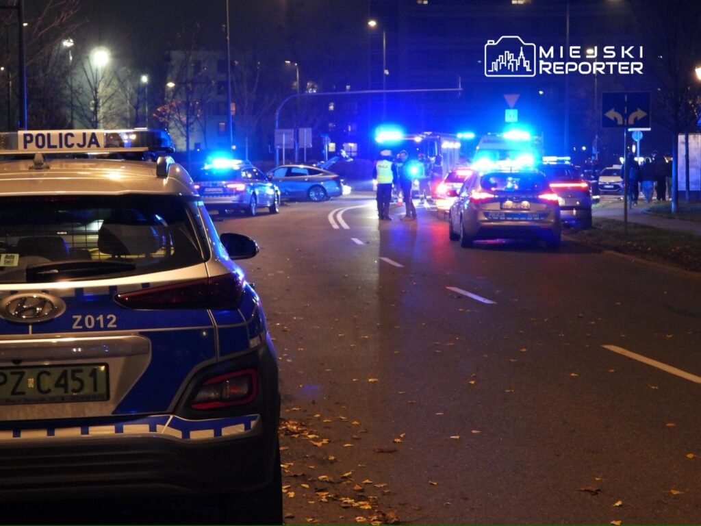 Policyjny radiowóz z niebieskimi światłami blokuje drogę, a funkcjonariusze kontrolują ruch na ulicy w nocy.