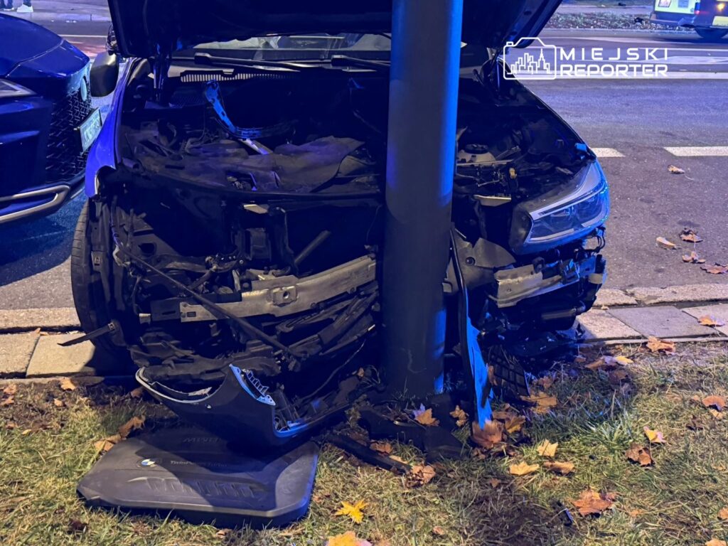 Zniszczony samochód osobowy uderzony w latarnię, z otwartą maską i uszkodzoną przednią częścią, na miejskiej ulicy.