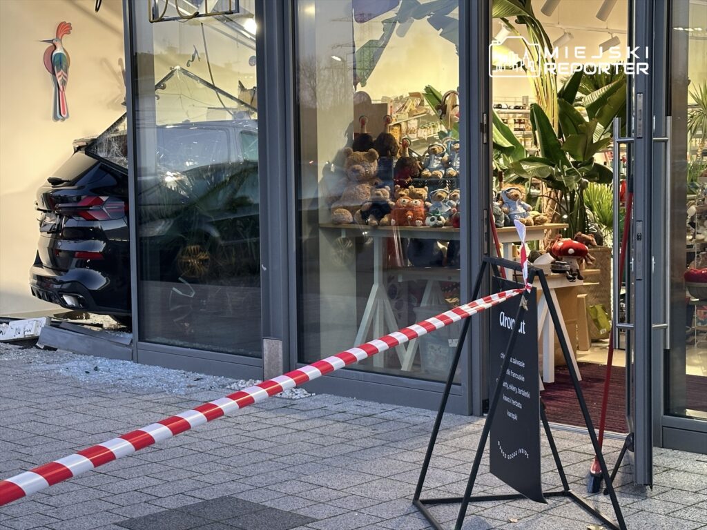 Czarny SUV wjeżdża przez szklane drzwi do sklepu z pluszakami, otoczenie zabezpieczone taśmą.