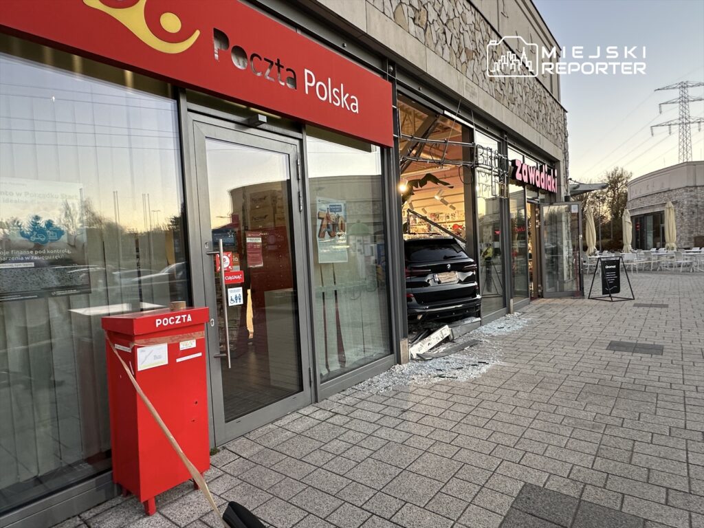 Samochód osobowy wjeżdża do sklepu spożywczego, rozbijając szklane drzwi, obok znajduje się skrzynka pocztowa.