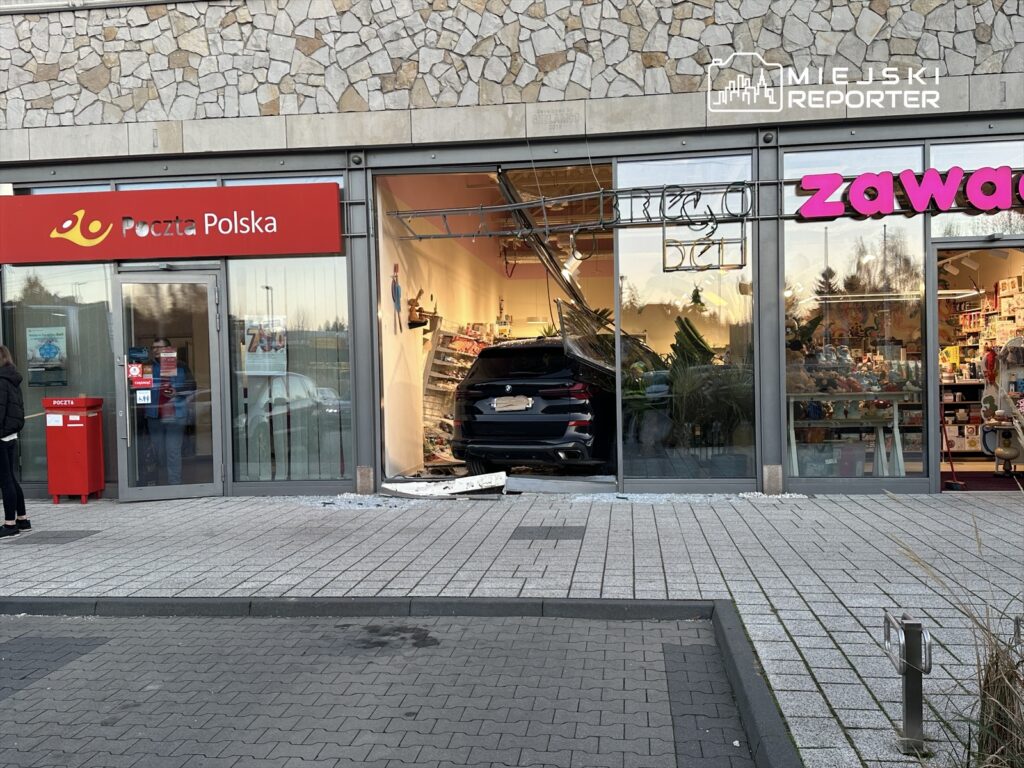 Czarny samochód wjeżdża przez witrynę sklepu, zniszczając część ściany i rozrzucając szkło na chodniku przed pocztą.
