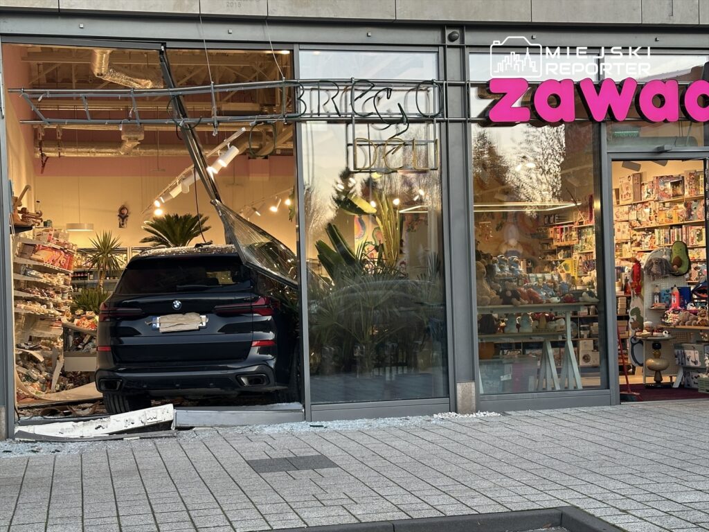 Czarny SUV wjeżdża przez szklane drzwi do sklepu z zabawkami, rozbijając witrynę i pozostawiając szkło na podłodze.