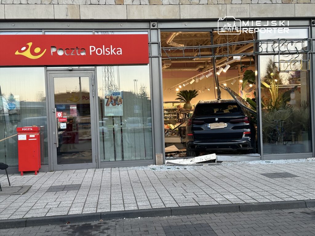 Czarny samochód osobowy wjeżdża przez przeszkloną witrynę poczty, zniszczone szkło leży na chodniku.
