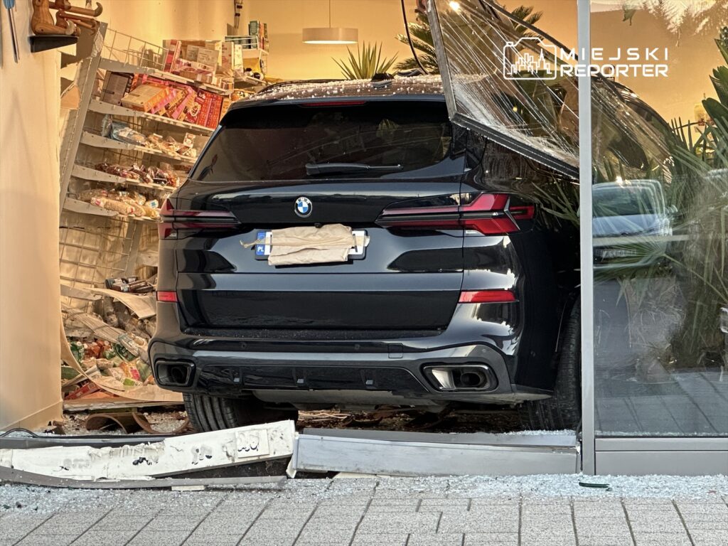 Czarny SUV wjeżdża przez przeszkloną ścianę sklepu, uszkadzając półki z towarami i rozrzucając produkty na podłodze.