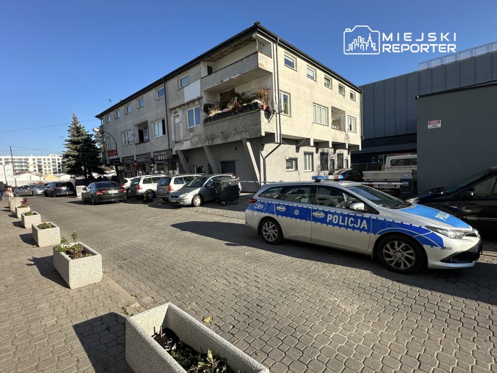 Policyjny radiowóz zaparkowany na chodniku wzdłuż ulicy, obok budynku mieszkalnego z lokalami usługowymi.