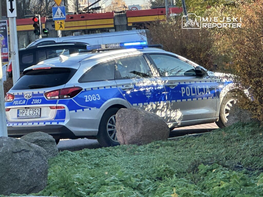 Policyjny radiowóz z niebieskimi światłami zatrzymany przy sygnalizacji świetlnej na skrzyżowaniu.