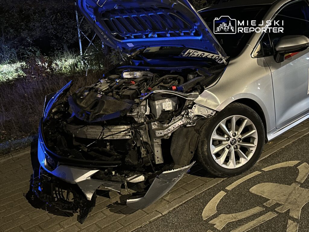 Uszkodzony samochód osobowy z otwartą maską, stojący na poboczu drogi w nocy. Z przodu widoczne uszkodzenia nadwozia.