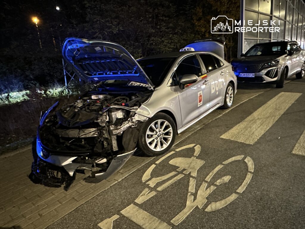 Zniszczony samochód osobowy z otwartą maską stoi na chodniku obok innych pojazdów w nocy.