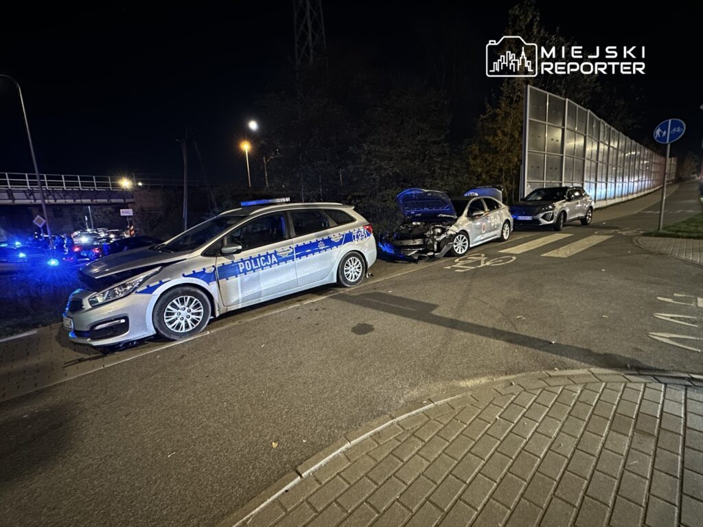 Policyjny samochód zaparkowany obok uszkodzonego pojazdu na ulicy, w tle widoczne niebieskie światła radiowozów.
