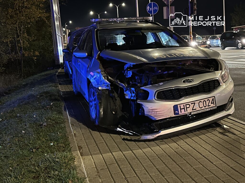 Uszkodzony radiowóz policyjny zaparkowany na chodniku, z widocznymi uszkodzeniami z przodu, w nocy.