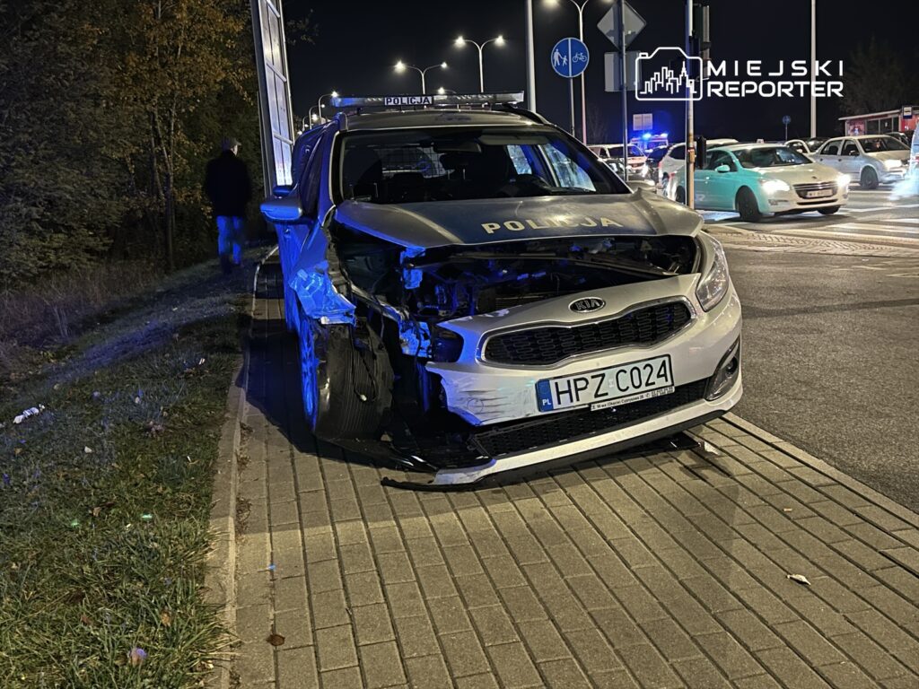 Uszkodzony radiowóz policyjny z wgniecionym przodem stoi na poboczu w nocy, obok przejazdu drogowego.