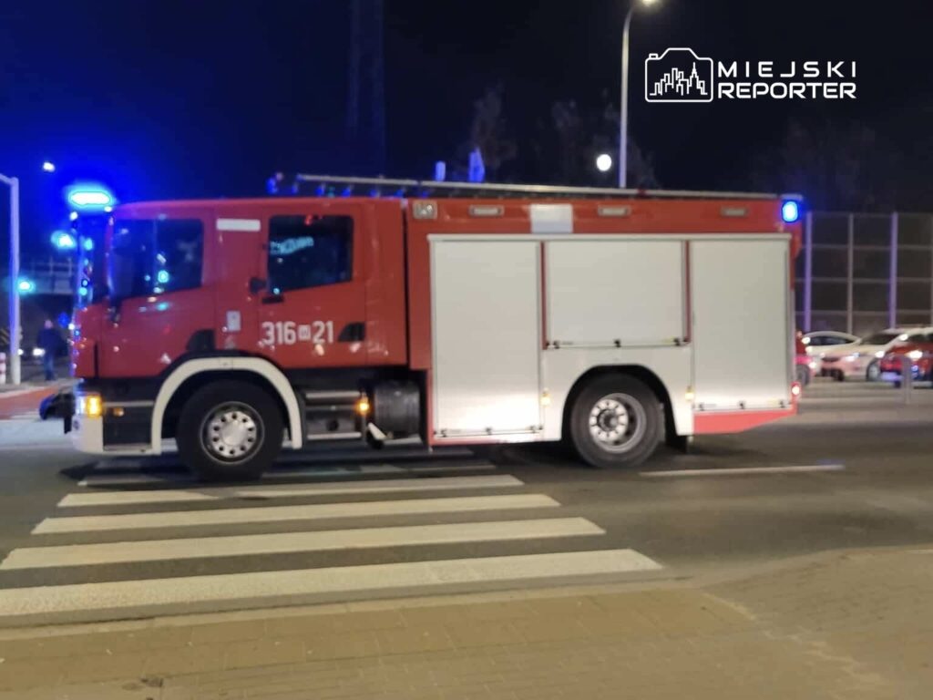 Czerwony wóz strażacki przejeżdża przez przejście dla pieszych w nocy, z włączonymi sygnałami świetlnymi.