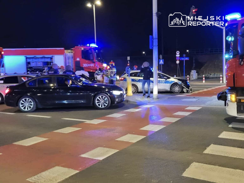 Na skrzyżowaniu nocą, czarne auto stoi obok uszkodzonego pojazdu policji, a ratownicy i policjanci prowadzą działania.