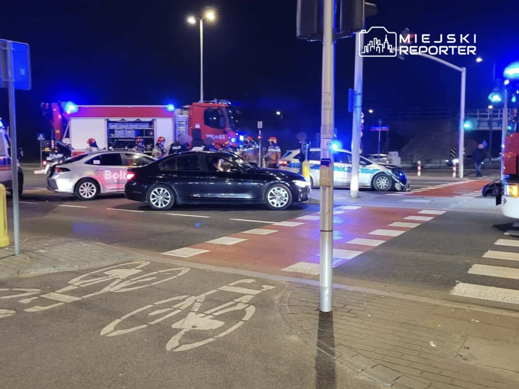 Na skrzyżowaniu z widocznymi pojazdami ratunkowymi, policjanci zabezpieczają miejsce zdarzenia w nocy.