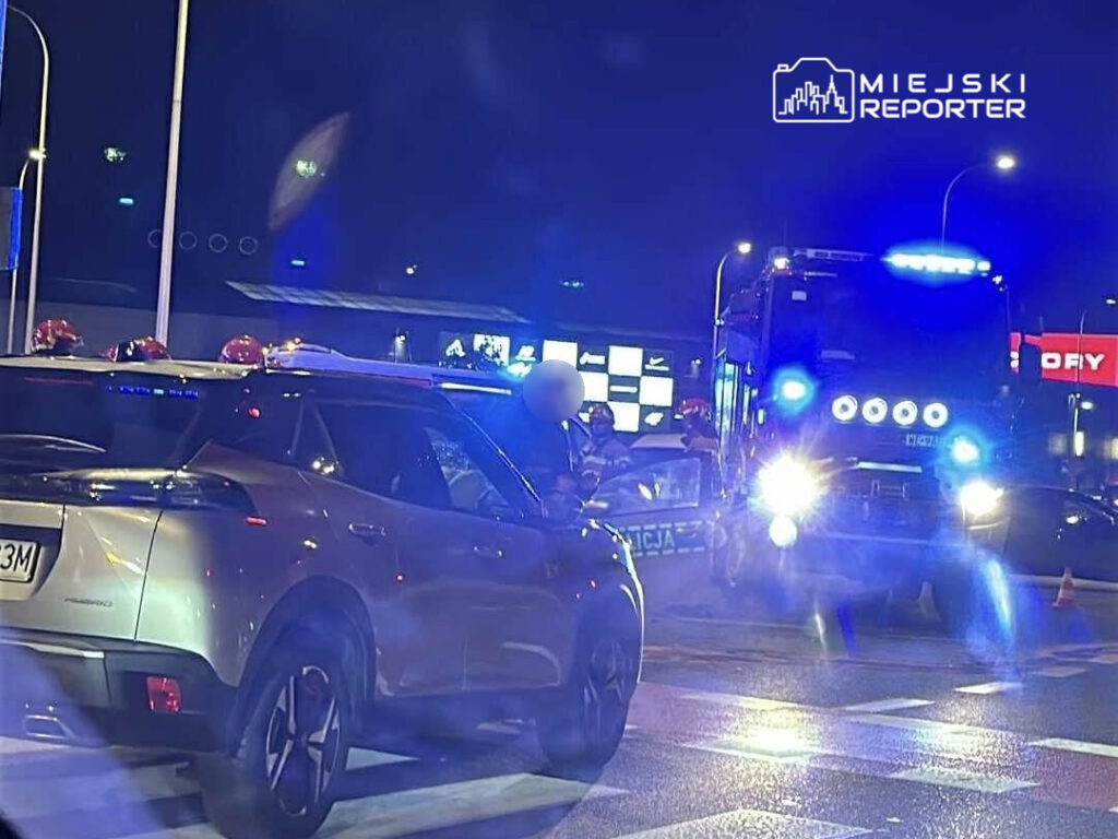 Policyjny radiowóz z włączonymi sygnałami świetlnymi stoi na skrzyżowaniu, obok srebrny samochód osobowy.