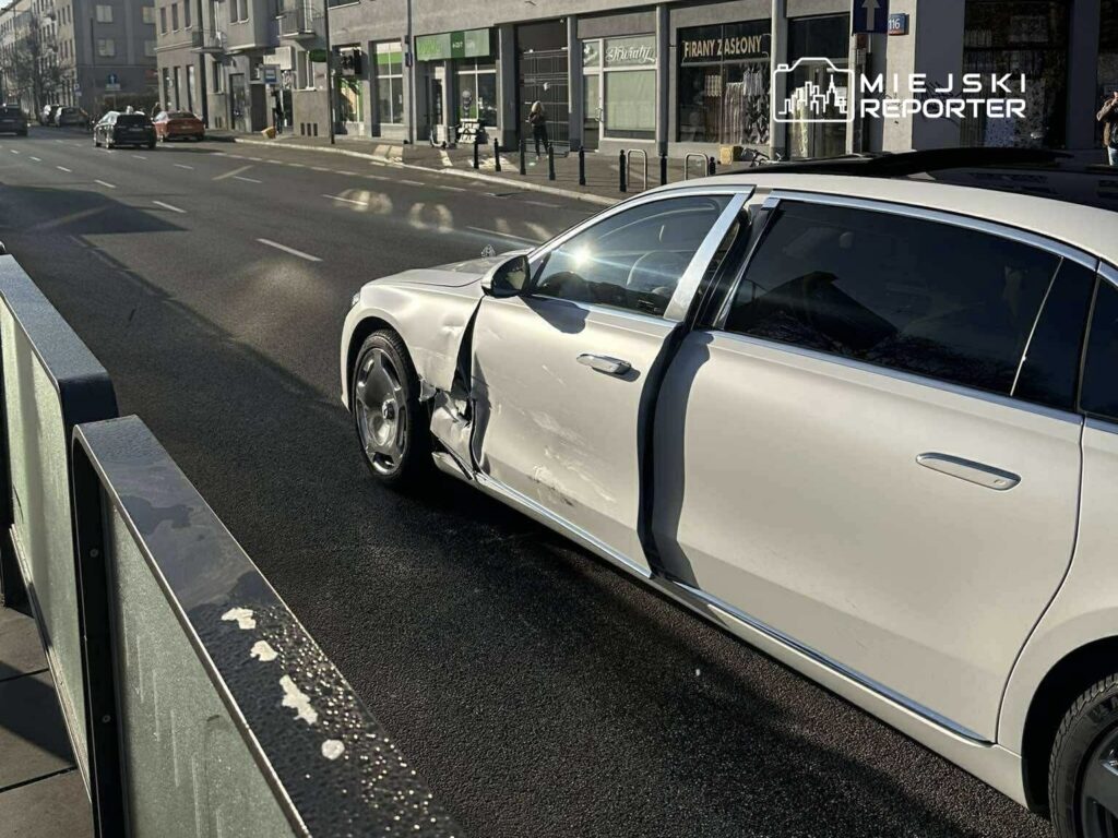 Uszkodzony biały samochód osobowy zaparkowany na ulicy, z otwartymi drzwiami po stronie kierowcy.