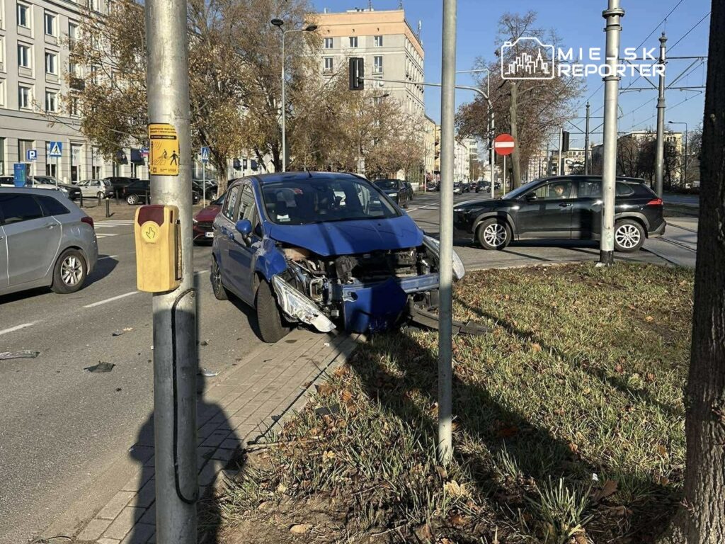 Niebieski samochód osobowy z uszkodzoną przednią częścią stoi obok słupa sygnalizacyjnego na ruchliwej ulicy.