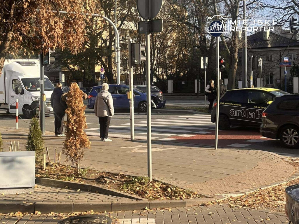 Grupa pieszych czeka na zielone światło na przejściu dla pieszych, obok ruchliwej ulicy z samochodami.