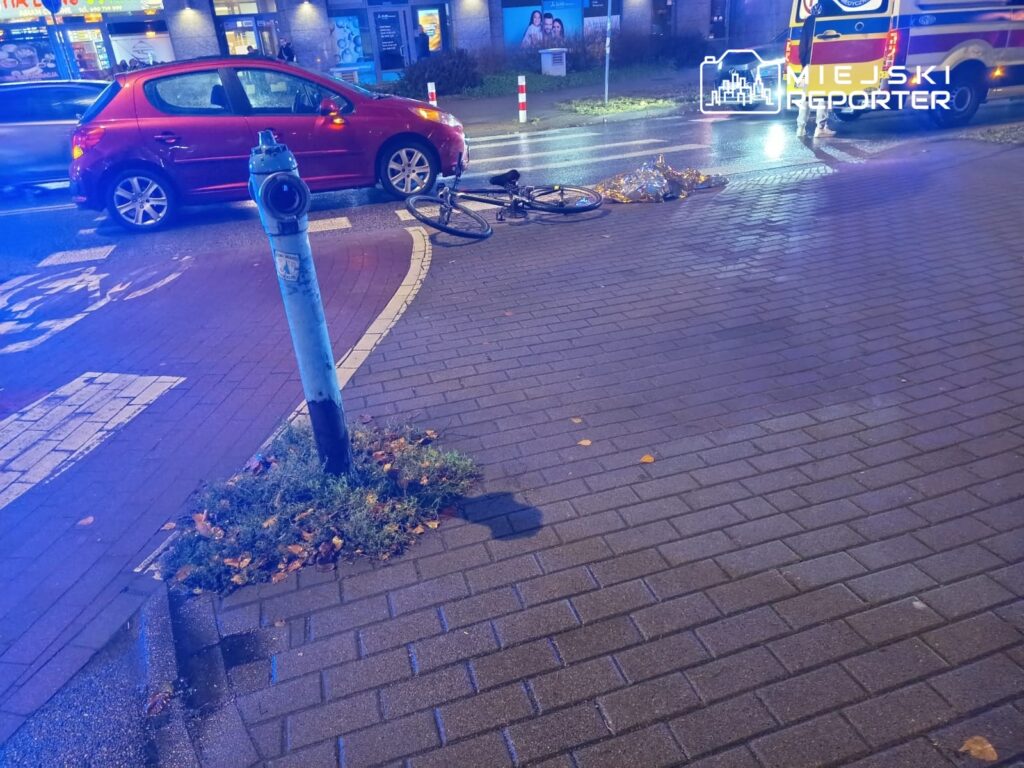 Na jezdni leży przewrócony rower obok hydrantu, a w tle widoczne są zaparkowane samochody i karetka.