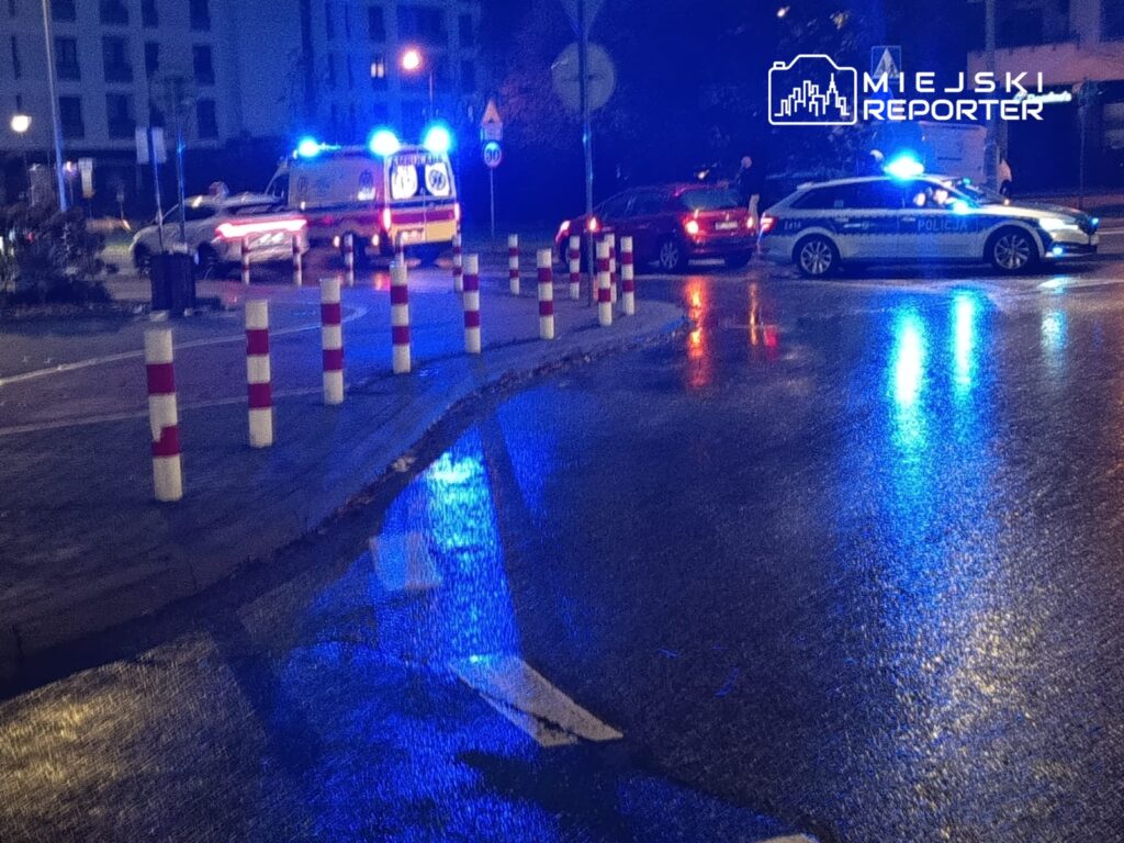 Na mokrej nawierzchni ulicy stoją samochody policyjne i karetka, a w tle widoczne są sygnały świetlne.