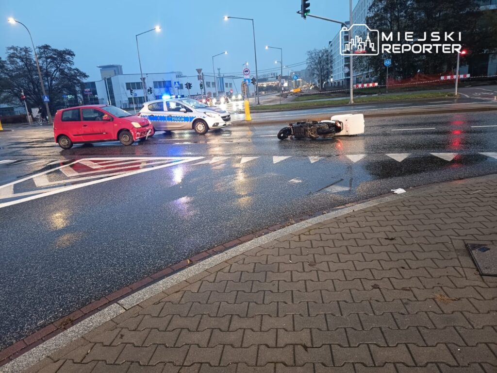 Czerwony samochód osobowy z uszkodzeniami stoi obok przewróconego skutera na mokrej nawierzchni drogi, w tle radiowóz policyjny.