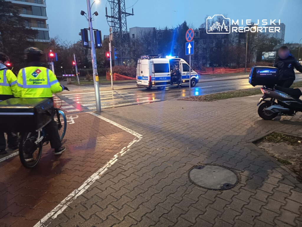 Dwaj policjanci w żółtych kurtkach stoją obok roweru dostawczego, podczas gdy policyjny van zatrzymał się na skrzyżowaniu.